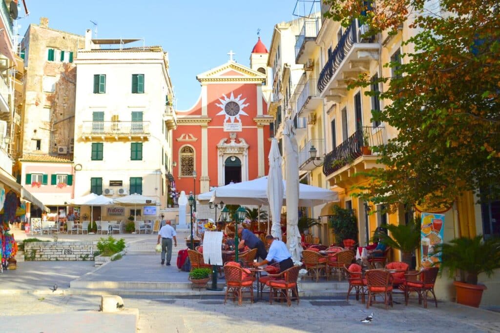 A cobbled square lined with outdoor cafes surrounded by pastel neoclassical buildings on Corfu Greece